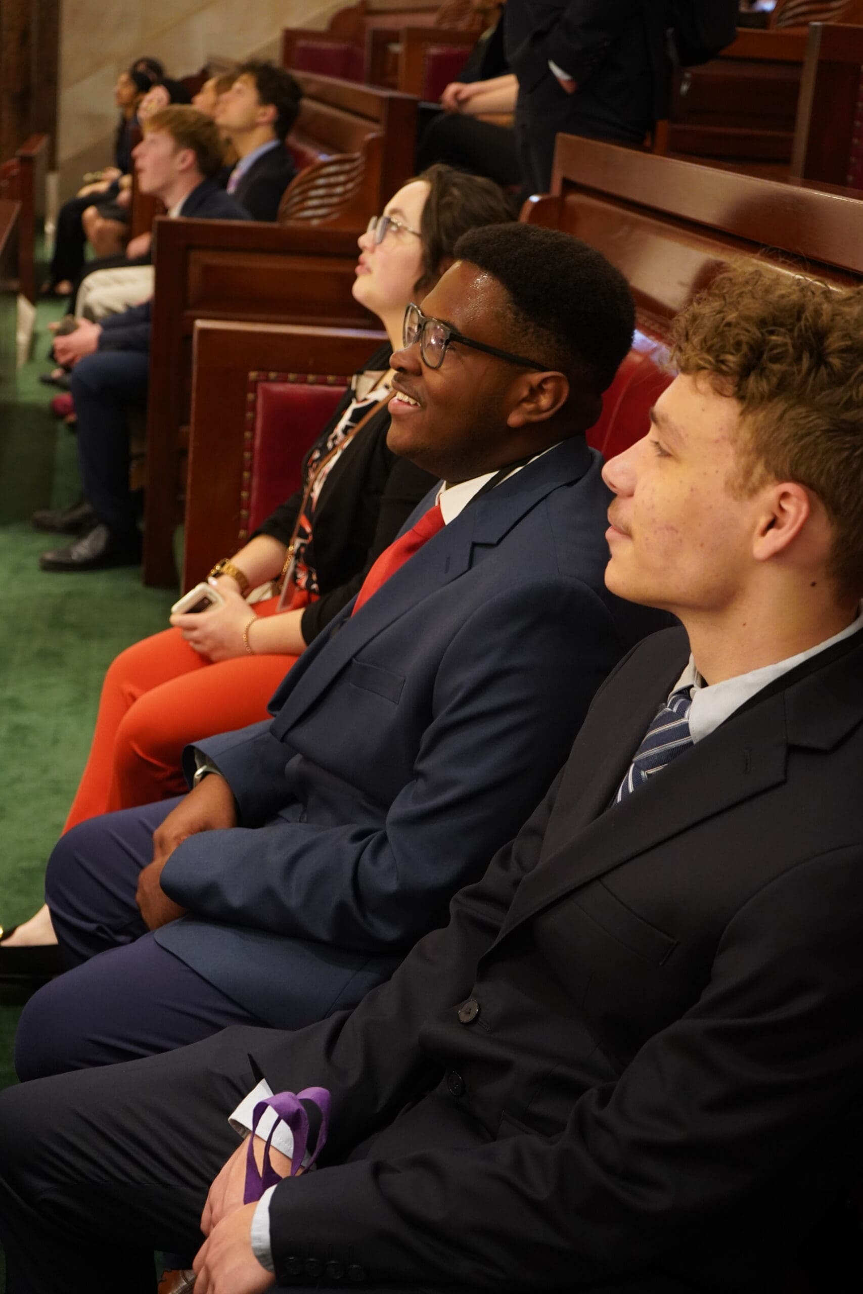 People in formal attire sit attentively in wooden pews, likely at an event or ceremony.
