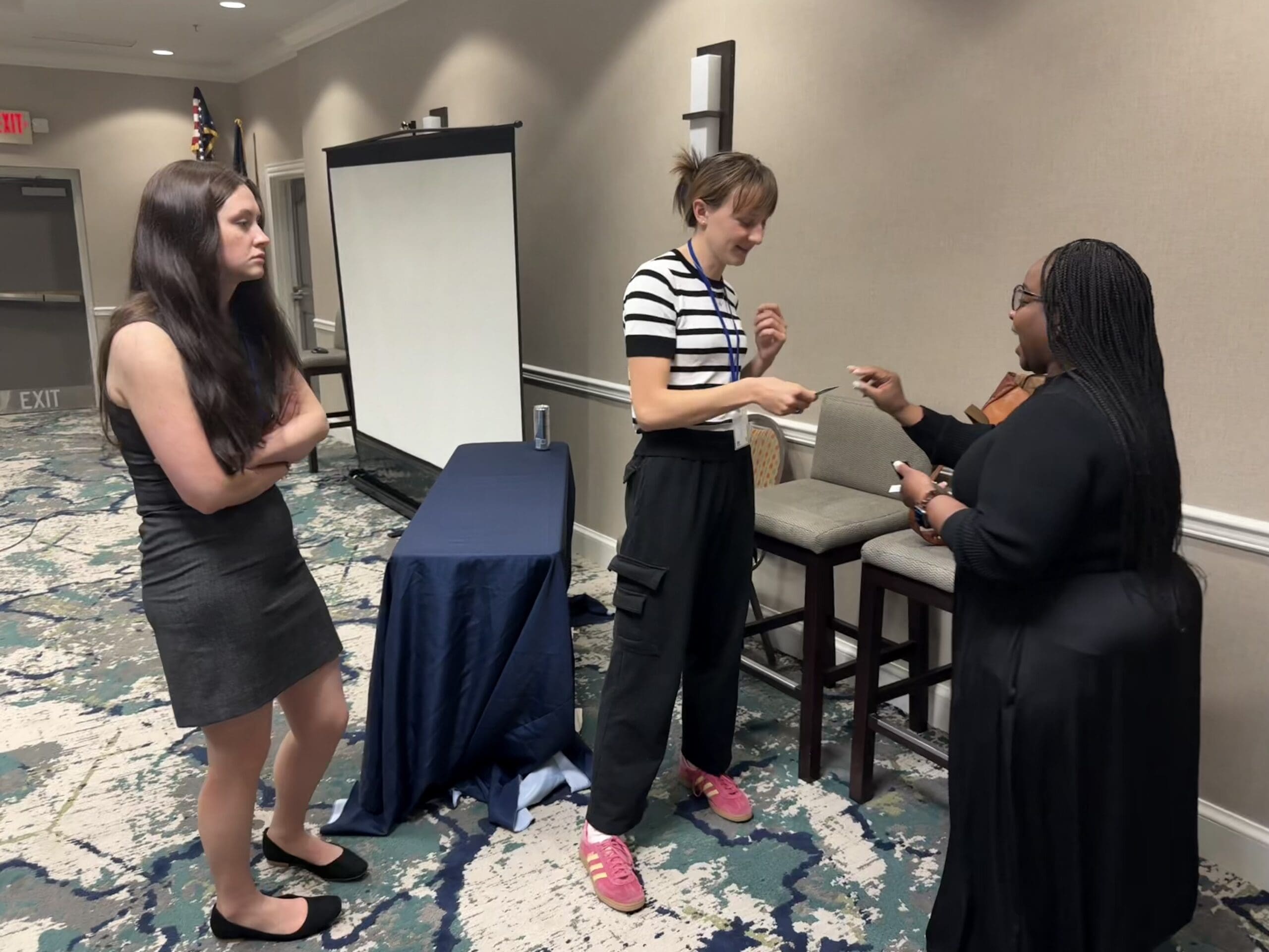 Three women stand in conversation near a table with a folded cloth and a projection screen in a conference room. One woman is handing an object to another.