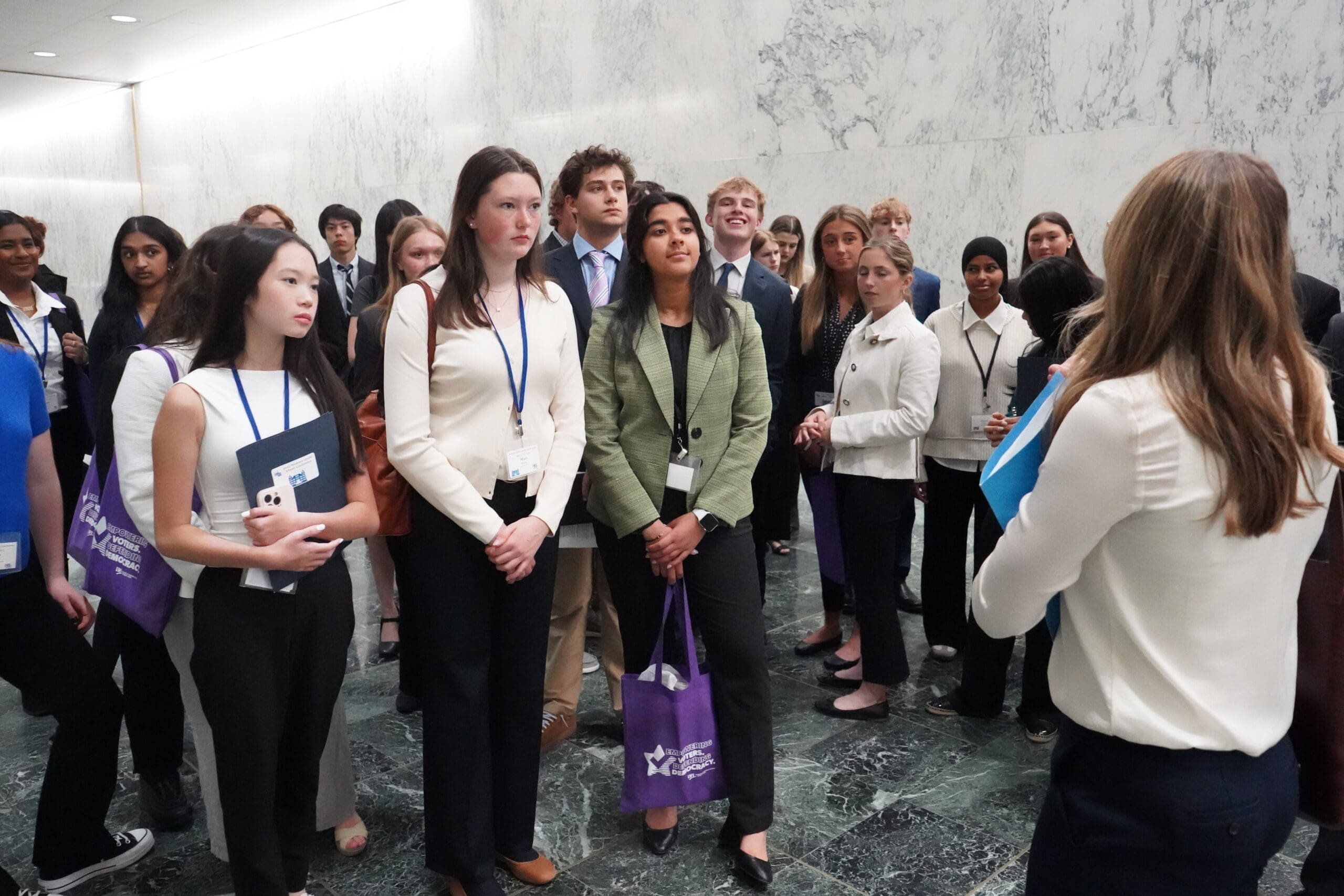 A group of young people, some holding folders and wearing name badges, listen to a woman speaking in a marble-walled hallway.