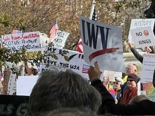 A crowd of people holding various protest signs, some with American flags, gathered outdoors amid trees.