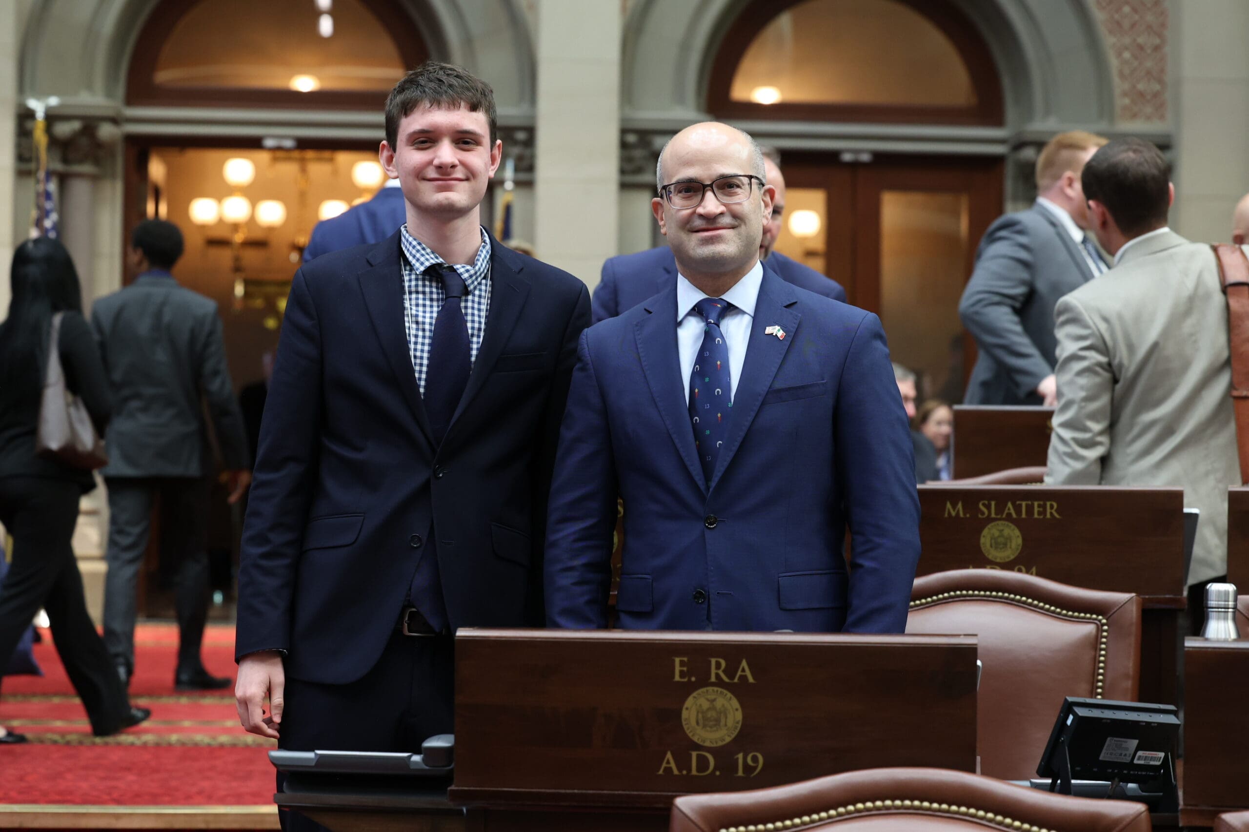 Two men in suits stand and pose for a photo inside a government chamber, in front of desks labeled "E. RA" and "M. SLATER." Other people are seen in the background.