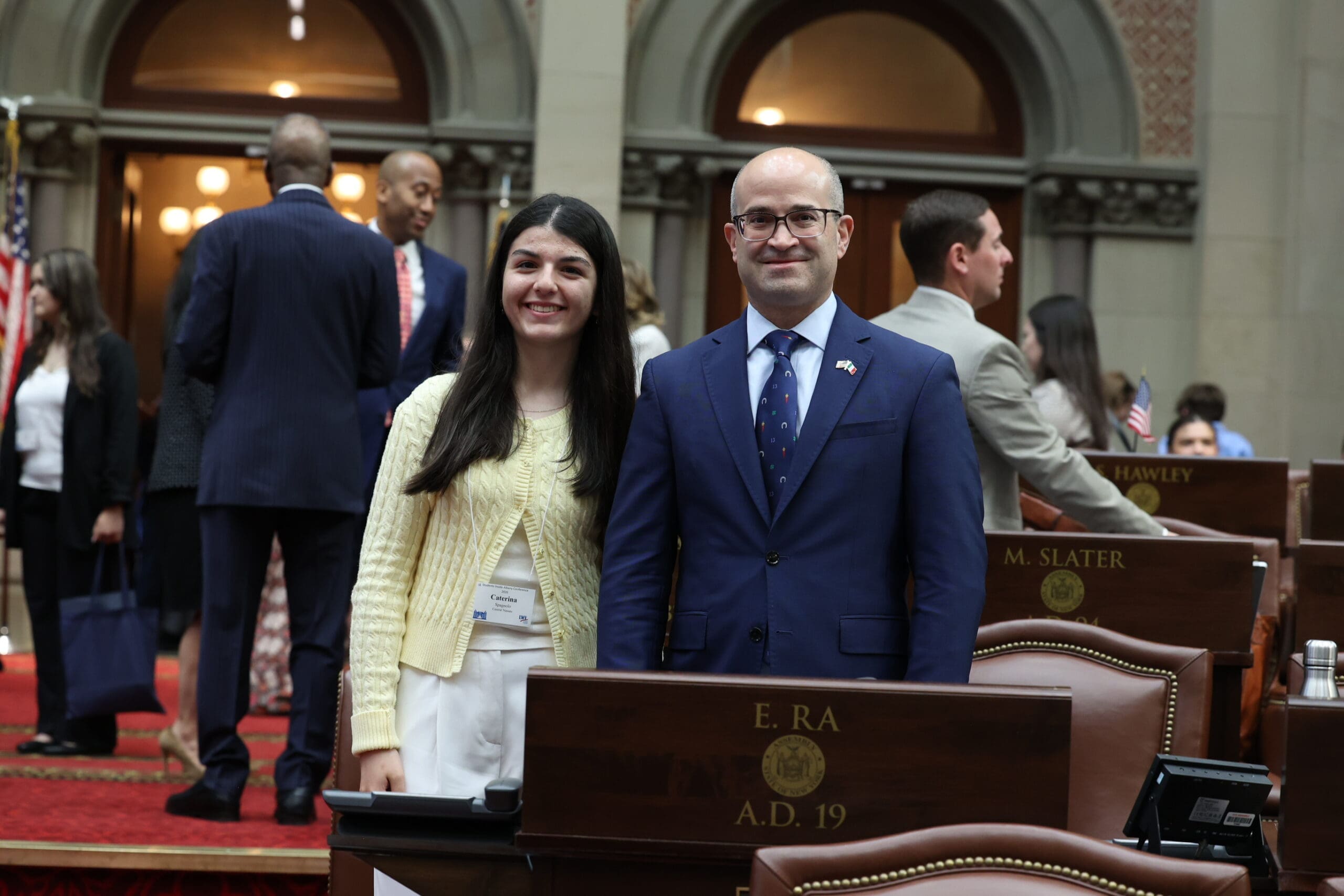 Two people stand and pose for a photo in a legislative chamber, with other individuals and desks visible in the background.