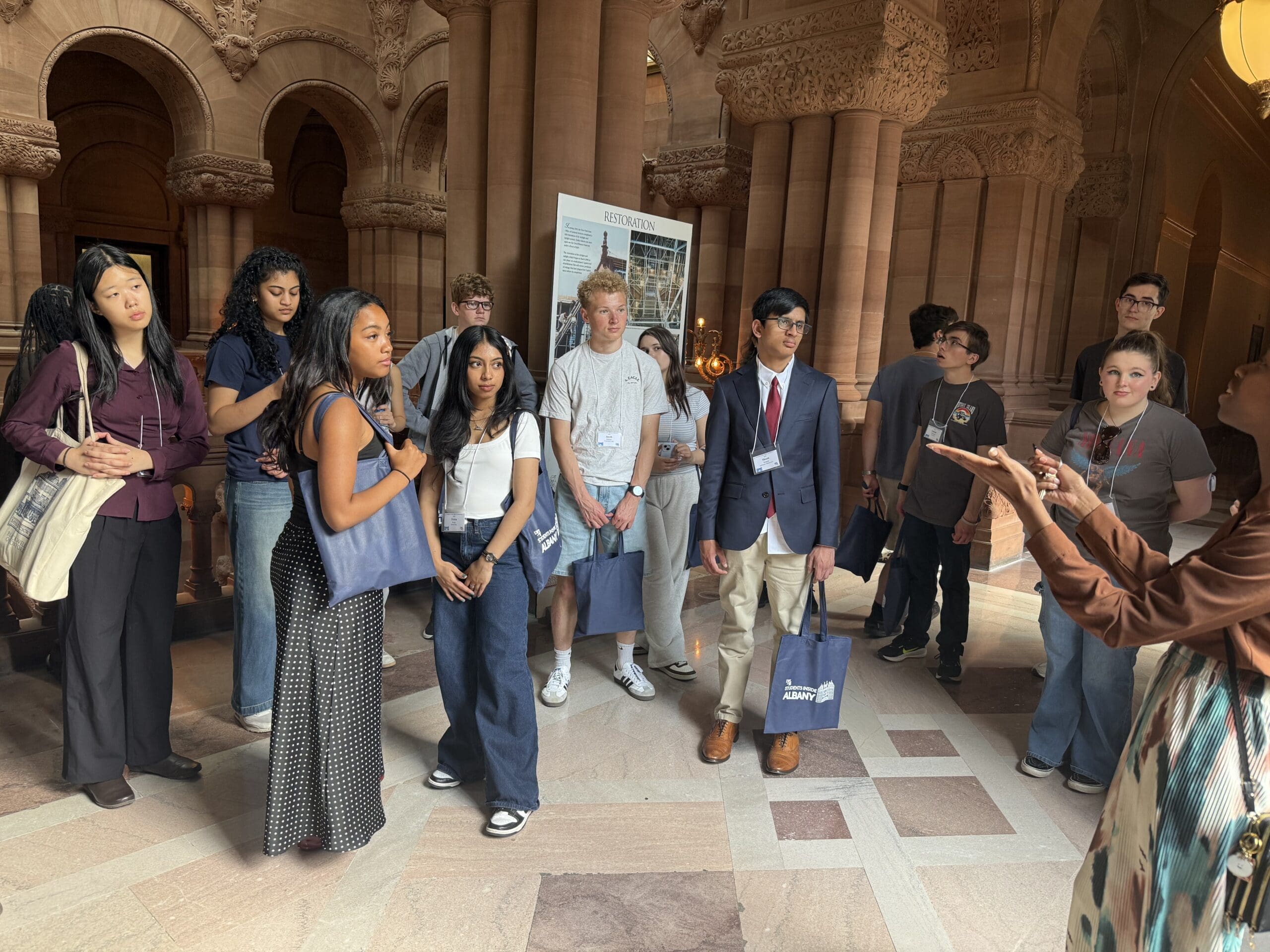 A group of students stand indoors, listening to a tour guide speaking and gesturing; the setting features ornate stone architecture.