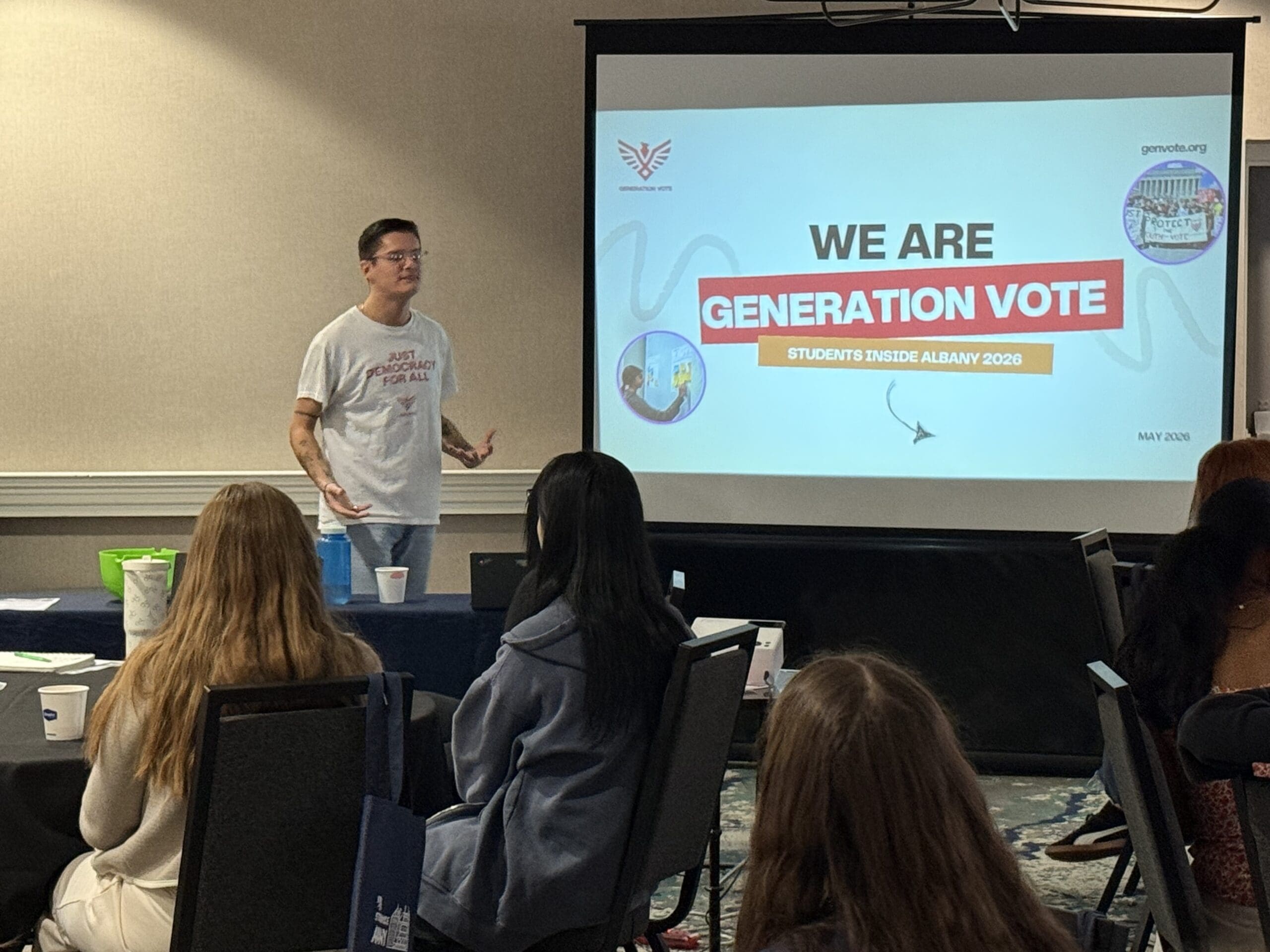 A speaker presents to a group of students in a conference room; the screen displays “We Are Generation Vote, Students Inside Albany 2023.”.