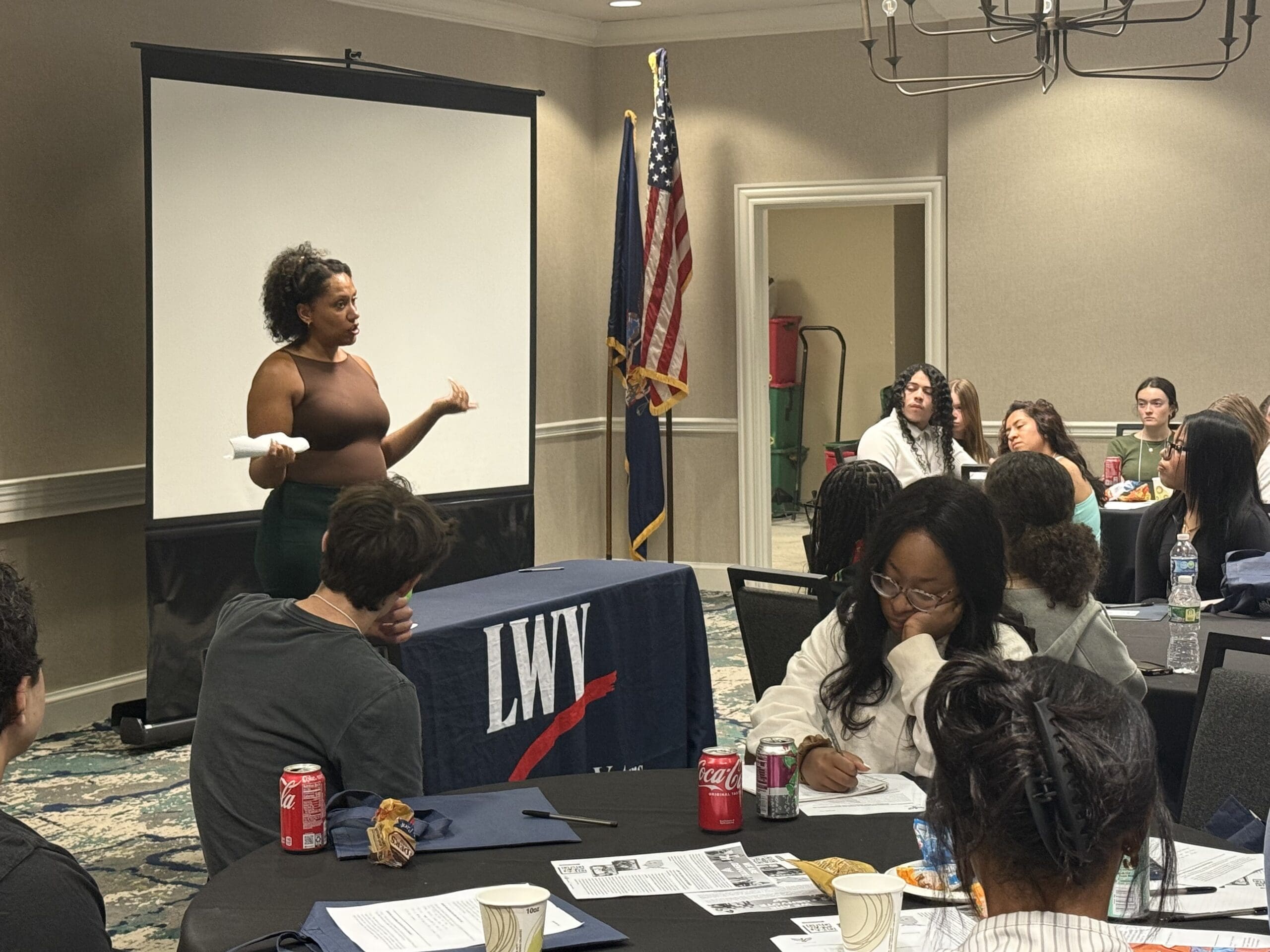 A woman speaks to a group seated around tables at a meeting. A screen, LWV table, and U.S. flag are visible in the room. Attendees are listening and taking notes.