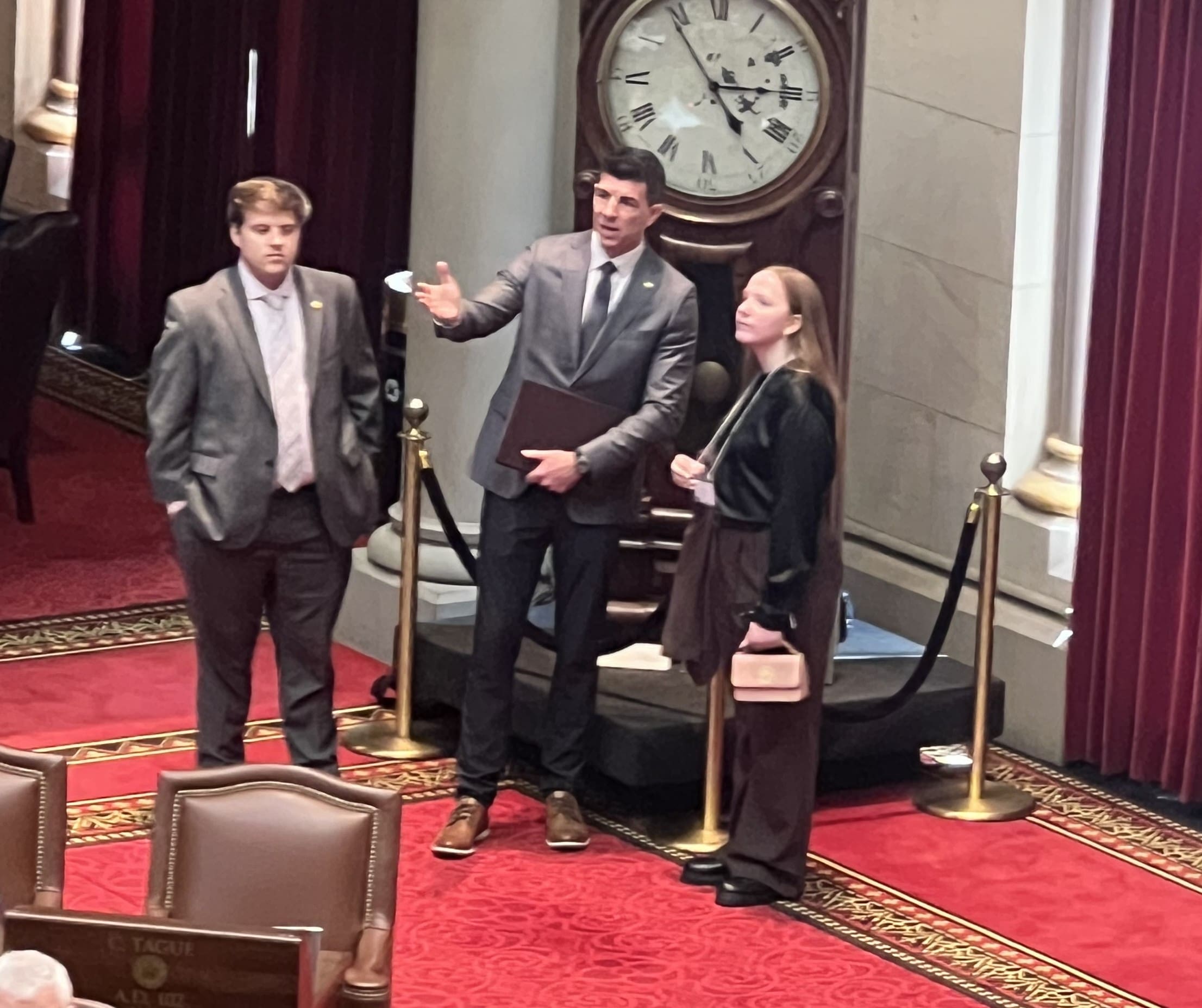 Three people in business attire stand talking in a formal room with red carpet, tall curtains, and a large wall clock in the background.