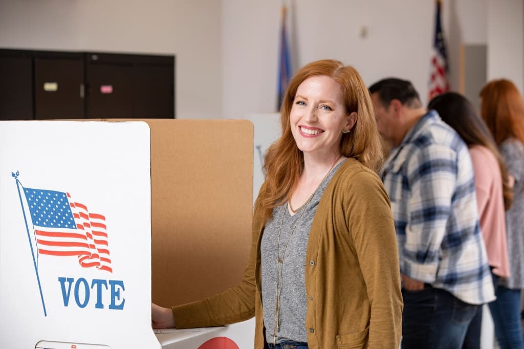 A woman stands at a voting booth, smiling at the camera, with other people lined up and voting in the background. An American flag and the word "VOTE" are visible on the booth.