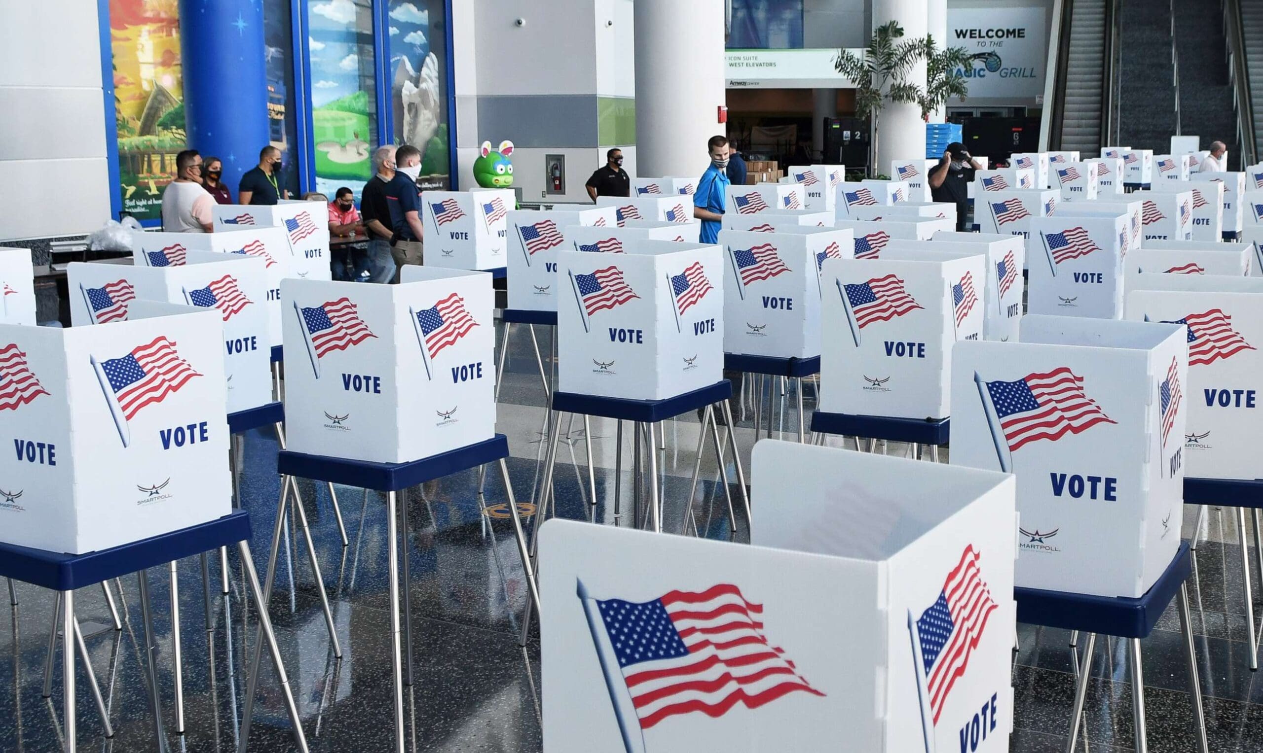 Rows of voting booths with American flags and "VOTE" signs are set up in a spacious indoor area, with people standing and sitting in the background.